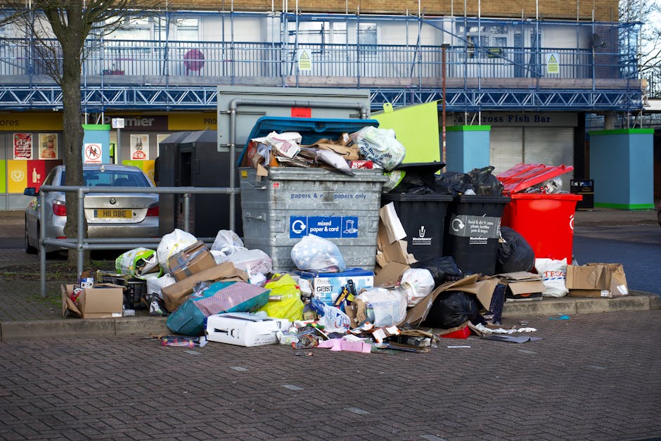 A person's hand is seen holding a green and blue plastic bin bag filled with waste, gripping the bag’s twisted top with fingers. The bag appears to contain household rubbish and is made of thin, sem